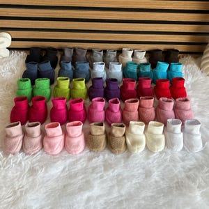 Könnte beinhalten: Eine Sammlung bunter Babyschuhe in verschiedenen Farbtönen, darunter Rosa, Blau, Grün und Braun. Die Schuhe sind auf einer flauschigen weißen Oberfläche angeordnet, mit einem Hintergrund aus Holzlatten.
