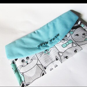 Könnte beinhalten: Eine Stoff-Clutch mit hellblauem Überschlag und einem Muster aus Cartoon-Katzen in Grau, Weiß und Türkis. Der Überschlag ist mit kleinen, runden, türkisfarbenen Perlen verziert. Die Clutch ist geschlossen.