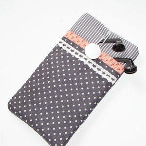 Op de afbeelding: Een grijze en witte polkadot telefoonhoes met een gestreepte bovenklep. De hoes heeft een witte kanten rand en een perzikkleurig accent. Een witte knop zet de klep vast en zwarte oortelefoons zijn zichtbaar.