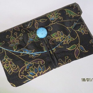 Könnte beinhalten: Eine schwarze Stofftasche mit Blumenmuster und einem blauen Knopfverschluss. Der Stoff hat einen goldenen Schimmer.