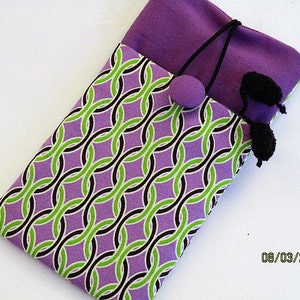 May include: Purple and green patterned fabric phone case with black pom-poms. The case is made of a soft, plush fabric and has a geometric design.