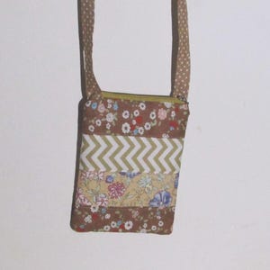 Op de afbeelding: Een kleine, rechthoekige crossbodytas met een bruin bloemenpatroon en een zigzagontwerp. De tas heeft een lange, gestippelde riem en een ritssluiting. De tas is gemaakt van stof en is geschikt voor kleine spullen.