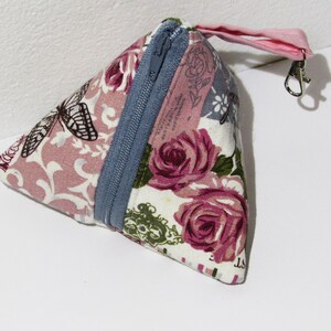 Può includere: Un astuccio triangolare in tessuto con un motivo floreale e a farfalle. L'astuccio presenta una cerniera blu e un cinturino rosa con un fermaglio in metallo. Il tessuto presenta rose rosa, farfalle e uno sfondo rosa chiaro.