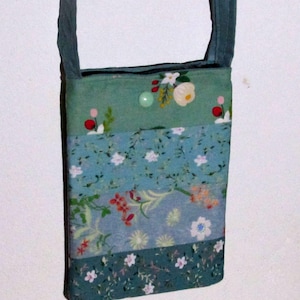 Könnte beinhalten: Handgefertigte, rechteckige Umhängetasche mit dunkelgrünem Riemen. Die Tasche besteht aus mehreren Stoffbahnen in Grün- und Blautönen, verziert mit floralen Mustern und gestickten Details. Ein grüner Knopf sichert die Oberseite.