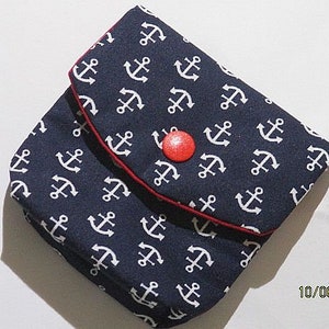 Könnte beinhalten: Eine kleine Stofftasche mit dunkelblauem Hintergrund und einem sich wiederholenden weißen Anker-Muster. Die Klappe wird mit einem einzelnen leuchtend roten runden Knopf verschlossen und hat einen dünnen roten Stoffrand an der Kante.