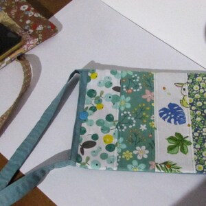 Könnte beinhalten: Eine rechteckige Umhängetasche mit floralem Muster, einem tealfarbenen Riemen und einem blauen Knopfverschluss. Die Tasche hat ein Patchwork-Design mit verschiedenen Blumen- und Blättermustern in Grün-, Weiß- und Rosatönen. Im Hintergrund ist ein Smartphone zu sehen.