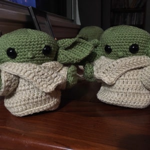 May include: Handmade crochet figures of a character with large ears and big black eyes. The figures are green with tan-colored clothing. The figures are displayed on a wooden surface. The figures are inspired by a popular science fiction character.