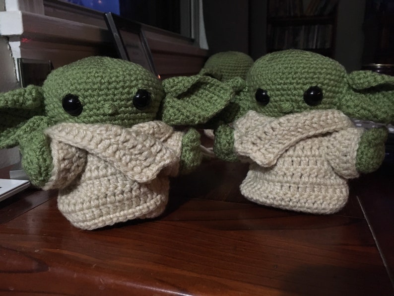 May include: Handmade crochet figures of a character with large ears and big black eyes. The figures are green with tan-colored clothing. The figures are displayed on a wooden surface. The figures are inspired by a popular science fiction character.