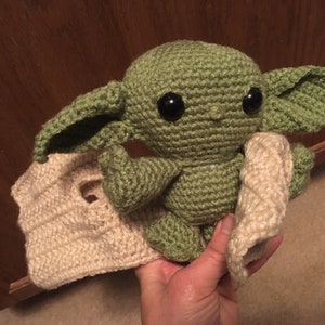 May include: A handmade green crocheted doll with large black eyes and oversized ears, resembling a character from a popular science fiction series. The doll is holding a cream-colored crocheted blanket. The doll is approximately 8 inches tall.