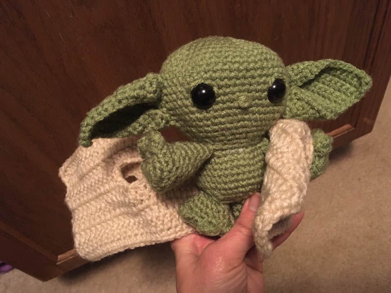 May include: A handmade green crocheted doll with large black eyes and oversized ears, resembling a character from a popular science fiction series. The doll is holding a cream-colored crocheted blanket. The doll is approximately 8 inches tall.