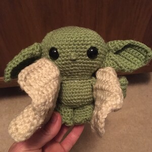 May include: A handmade crochet doll in the likeness of a character from a popular science fiction series. The doll is primarily green with large black eyes and cream-colored accents. The doll has large ears and is held in a hand.