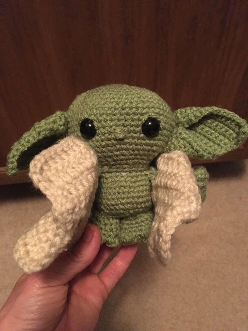 May include: A handmade crochet doll in the likeness of a character from a popular science fiction series. The doll is primarily green with large black eyes and cream-colored accents. The doll has large ears and is held in a hand.