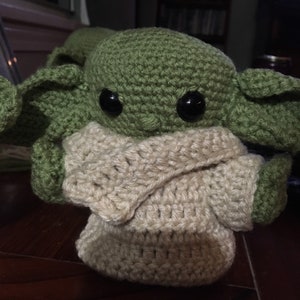 May include: A handmade crochet doll of a character with large black eyes, green skin, and large ears. The doll is wearing a cream-colored outfit. The doll is sitting on a wooden surface.