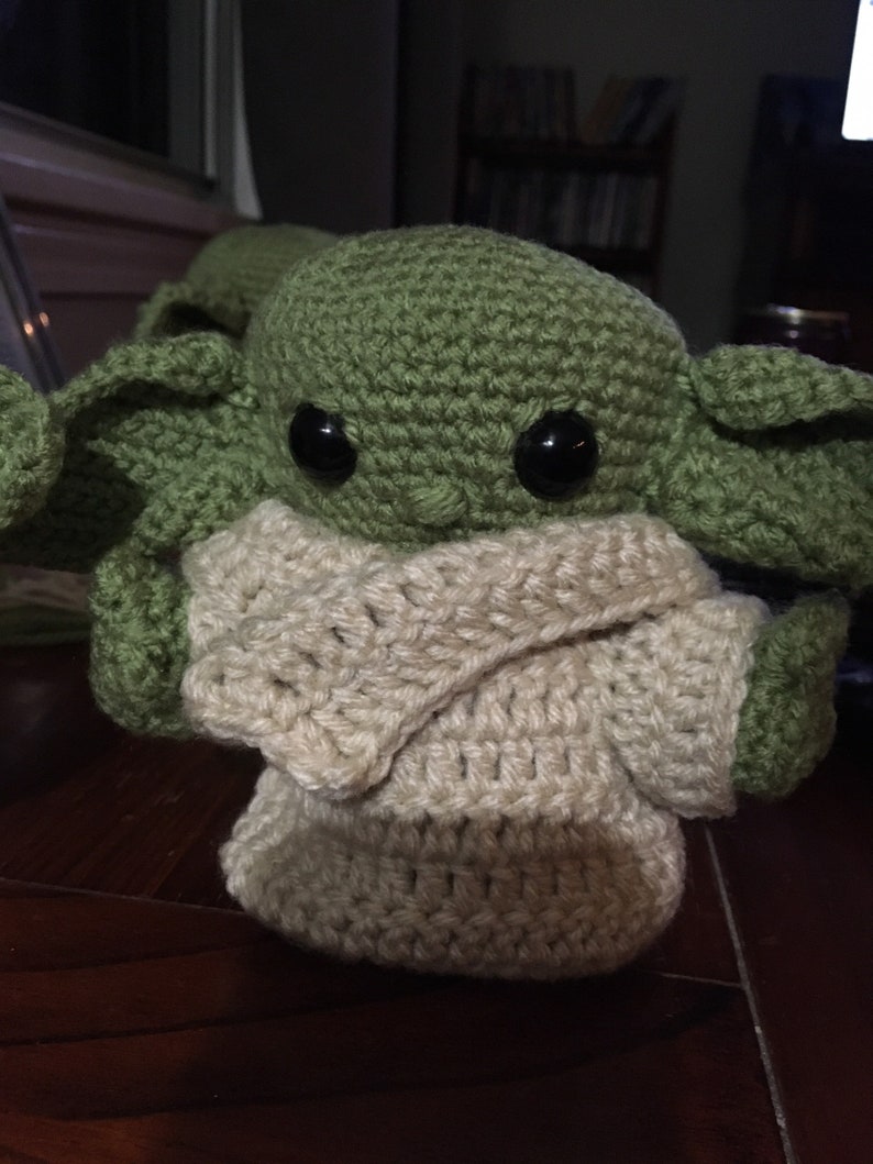 May include: A handmade crochet doll of a character with large black eyes, green skin, and large ears. The doll is wearing a cream-colored outfit. The doll is sitting on a wooden surface.