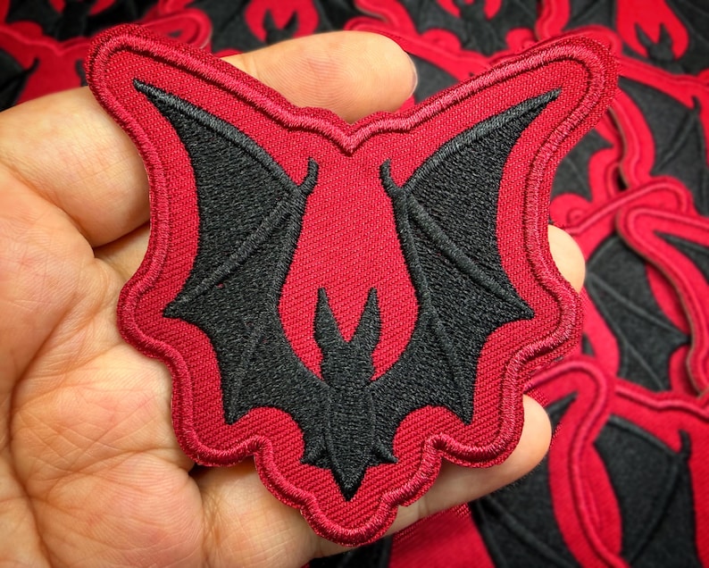 Peut inclure: &Eacute;cusson brod&eacute; repr&eacute;sentant une chauve-souris noire aux ailes d&eacute;ploy&eacute;es sur fond rouge. Le motif de la chauve-souris est d&eacute;taill&eacute; avec des coutures noires, et l'&eacute;cusson a une bordure rouge. Pour v&ecirc;tements ou accessoires.