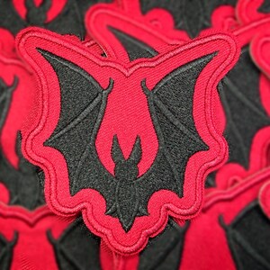 Peut inclure: &Eacute;cusson brod&eacute; avec un motif de chauve-souris noire sur fond rouge. La chauve-souris a des ailes d&eacute;ploy&eacute;es et est encadr&eacute;e d'une bordure rouge. Pour v&ecirc;tements ou accessoires.