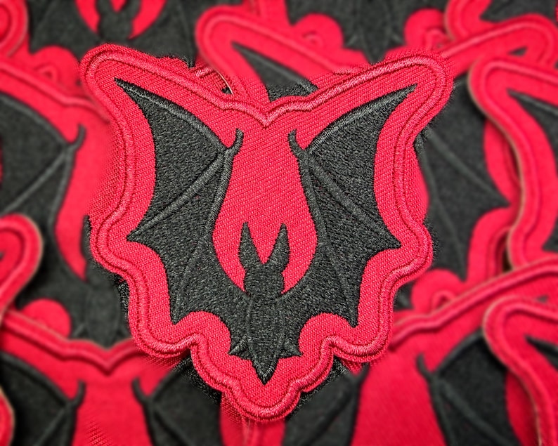 Peut inclure: &Eacute;cusson brod&eacute; avec un motif de chauve-souris noire sur fond rouge. La chauve-souris a des ailes d&eacute;ploy&eacute;es et est encadr&eacute;e d'une bordure rouge. Pour v&ecirc;tements ou accessoires.