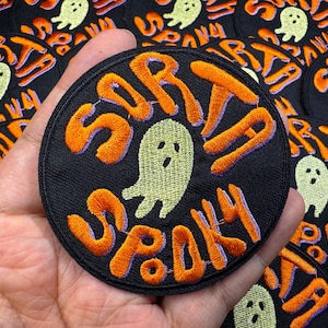 May include: Round black embroidered patch with the words "Sorta Spooky" in orange, outlined in purple. A light green ghost is in the centre. The patch is held in a hand, with other similar patches in the background.