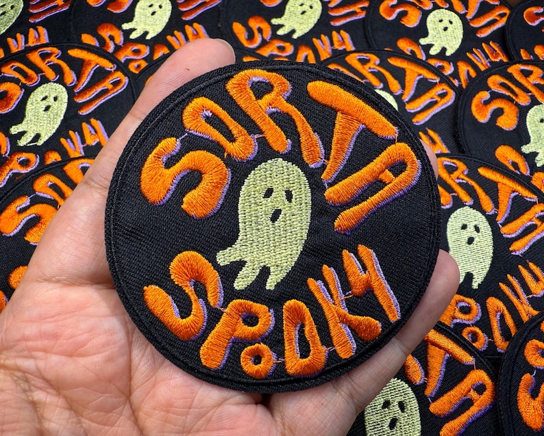以下が含まれることがあります： オレンジ色で紫色の縁取りが施された「Sorta Spooky」の文字が刺繍された、黒い丸いパッチ。中央には薄緑色の幽霊がいます。パッチは手で持たれており、背景には同様のパッチがいくつかあります。