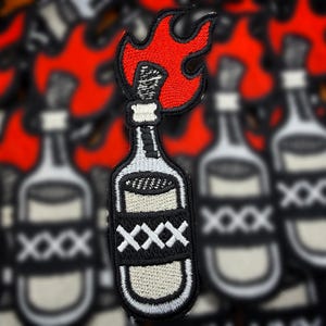 May include: An embroidered patch featuring a white bottle with a black label marked with three white X symbols. A bright red flame emerges from the top of the bottle neck, which is wrapped in a white cloth. The design has a thick black border.