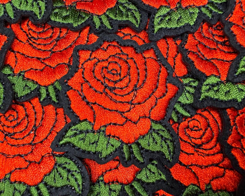 Puede incluir: Primer plano de parches bordados con rosas rojas con contornos negros y hojas verdes. Las rosas est&aacute;n densamente empaquetadas, creando un vibrante patr&oacute;n floral. Los parches son probablemente para fines decorativos.