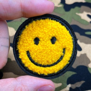 Peut inclure: Un patch smiley jaune avec un contour noir. Le patch est fait de tissu pelucheux et mesure environ 5 cm de diamètre.