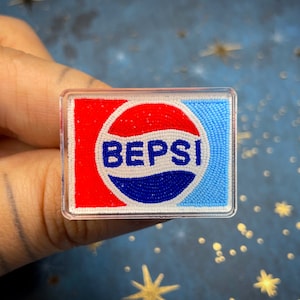 May include: A rectangular pin featuring the "BEPSI" logo, a parody of the Pepsi logo. The design includes a red and blue colour scheme with white accents. The pin is encased in a clear, rectangular frame.