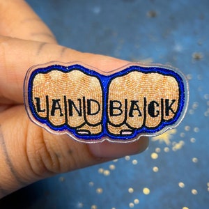 Peut inclure: Épinglette en acrylique transparent représentant deux poings avec les mots "LAND BACK" en lettres majuscules noires. Les poings sont de couleur beige avec un motif en mosaïque, soulignés en bleu et noir.