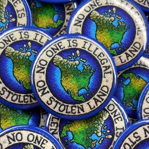 Puede incluir: Primer plano de varios botones redondos azules y blancos con el texto "No One Is Illegal On Stolen Land" y un mapa de mosaico de América del Norte.