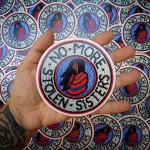 May include: A hand holds a round sticker with the text "No More Stolen Sisters" in a beaded design. The sticker is surrounded by other stickers with the same design.