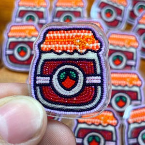 Puede incluir: Un broche de cuentas con forma de tarro de mermelada. El tarro es principalmente rojo con un borde blanco y morado. La tapa es naranja y el centro tiene un diseño verde y rojo. El broche se sostiene en una mano.