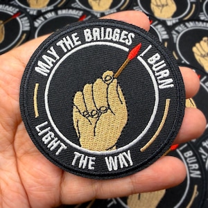 May include: Black round patch with the text "MAY THE BRIDGES I BURN LIGHT THE WAY". The patch features a hand holding a lit match. The hand and match are in shades of tan, red, and gold.