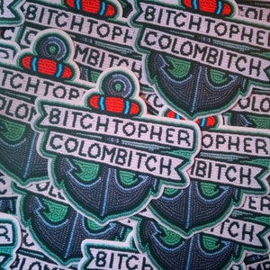 May include: A collection of embroidered patches with the text "BITCHTOPHER COLOMBITCH" in a banner over a navy blue anchor design. The patches have red, green, and white accents.
