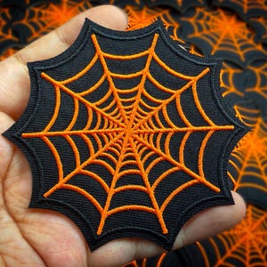 May include: A black, star-shaped patch with an orange spider web design. The web is finely embroidered, with lines radiating from the centre. This patch is likely for decorative use, perhaps for Halloween or craft projects.