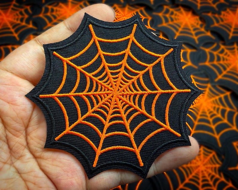 May include: A black, star-shaped patch with an orange spider web design. The web is finely embroidered, with lines radiating from the centre. This patch is likely for decorative use, perhaps for Halloween or craft projects.