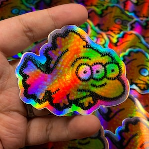 May include: Holographic sticker featuring a cartoon character with three eyes, outlined in black. The sticker has a rainbow color scheme with shades of orange, yellow, green, blue, and purple. The sticker is held in a hand, with other similar stickers in the background.