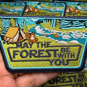 May include: Embroidered patch depicting a camping scene with a tent, campfire, and river. The patch has a green and blue colour scheme with the text "MAY THE FOREST BE WITH YOU" in a bold font. The patch is rectangular.