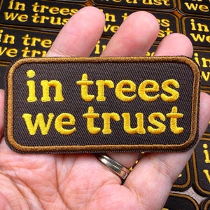 May include: A rectangular patch with the embroidered phrase "in trees we trust" in yellow letters on a dark brown background. The patch has a brown border and is held in a hand.