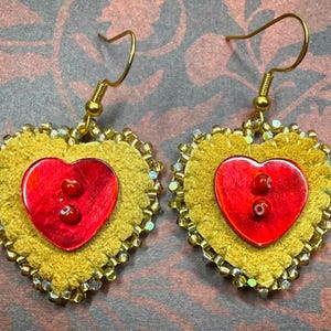 May include: A pair of heart-shaped earrings featuring a mustard yellow suede base with scalloped edges. Each earring is centred with a glossy red heart-shaped button containing two small red beads. The perimeter is lined with metallic gold beads.