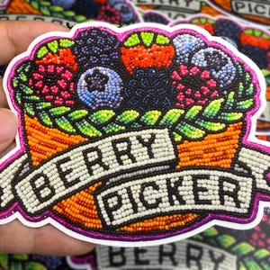 Może przedstawiać: Kolorowa naklejka przedstawiająca kosz pełen jagód w kolorach czerwonym, niebieskim i czarnym. Kosz jest pomarańczowy, a napis "BERRY PICKER" znajduje się na banerze poniżej. Biała obwódka.