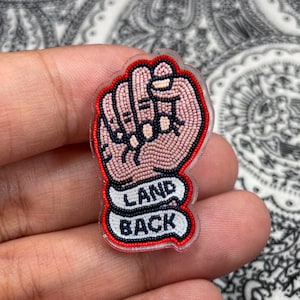 May include: A clear, plastic pin featuring a beaded fist in pink, black, and white. The fist is outlined in red and has the words "LAND BACK" in a white banner. The pin is likely a symbol of solidarity or activism.