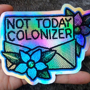 Könnte beinhalten: Holografischer Aufkleber mit dem Text "NOT TODAY COLONIZER" in Schwarz. Der Aufkleber hat die Form eines Umschlags mit Perlenblumen in Blau, Lila und Grün. Der Aufkleber hat einen Regenbogen-Schimmer.