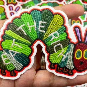 May include: A colorful embroidered patch of a caterpillar with the words "The Very Hungry Caterpillar" in green and white beads. The caterpillar is made of green, white, and red beads and has a red face with two black eyes.