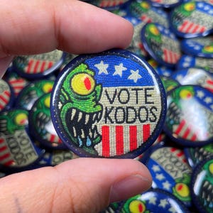 Puede incluir: Un botón redondo con borde azul y un diseño que presenta un monstruo verde con un ojo amarillo y el texto "VOTE KODOS". El botón también incluye rayas rojas y blancas y tres estrellas blancas.