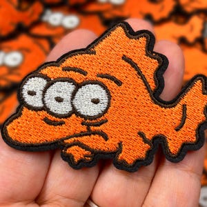 May include: Orange embroidered patch of a cartoon character with three eyes and a frown. The character has a textured, fuzzy appearance.