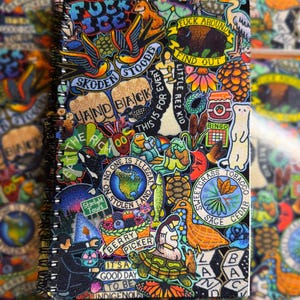 May include: A spiral-bound notebook featuring a dense collage of colourful illustrations and text, including phrases like FUCK ICE, FUCK AROUND FIND OUT, LAND BACK, NO ONE IS ILLEGAL ON STOLEN LAND, BERRY PICKER, and IT'S A GOOD DAY TO BE INDIGENOUS.