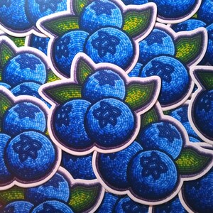 Op de afbeelding: Close-up van een vel stickers met meerdere blauwe bessenontwerpen. Elke blauwe bes is gemaakt van kleine mozaïekachtige stukjes in blauw, groen en wit. De stickers zijn gedrukt op wit papier.