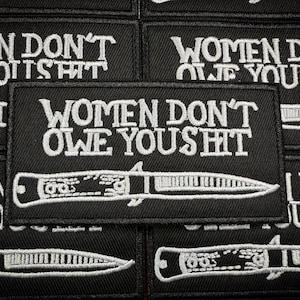 Op de afbeelding: Zwarte geborduurde patches met witte tekst die "WOMEN DON'T OWE YOU SHIT" en een mesontwerp weergeven. De patches zijn rechthoekig en hebben een geweven textuur.