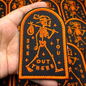 Peut inclure: Écusson brodé noir et orange représentant un squelette portant un sac sur un bâton. Le patch porte les mots "SEE YOU OUT THERE" en orange. Le patch est rectangulaire avec un sommet en arc et une bordure orange.