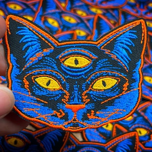 Pode incluir: Um remendo bordado azul e laranja de um gato com três olhos. Os olhos do gato são amarelos e o fundo é preto.